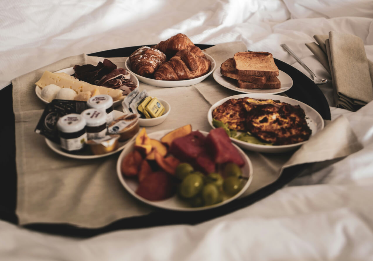 Breakfast tray on a white bedspread with croissants, cheese, charcuterie, butter, and assorted jams.