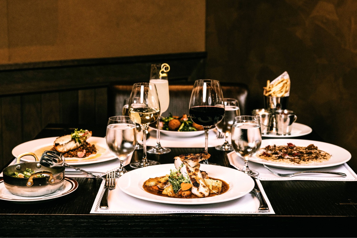 Fine dining tabletop with wine, cocktails and various entrees displayed ranging from fish to pork with roasted vegetables.